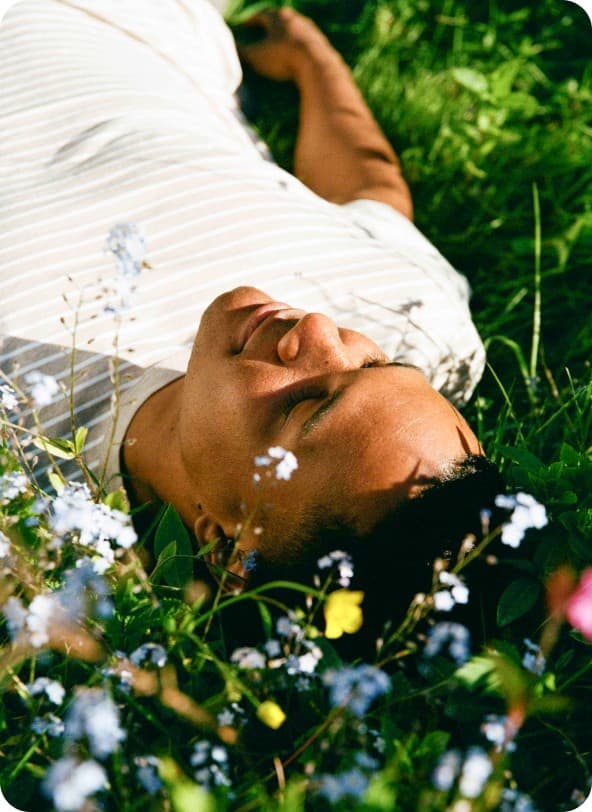 A young person resting on green grass among small flowers, eyes closed in warm sunlight